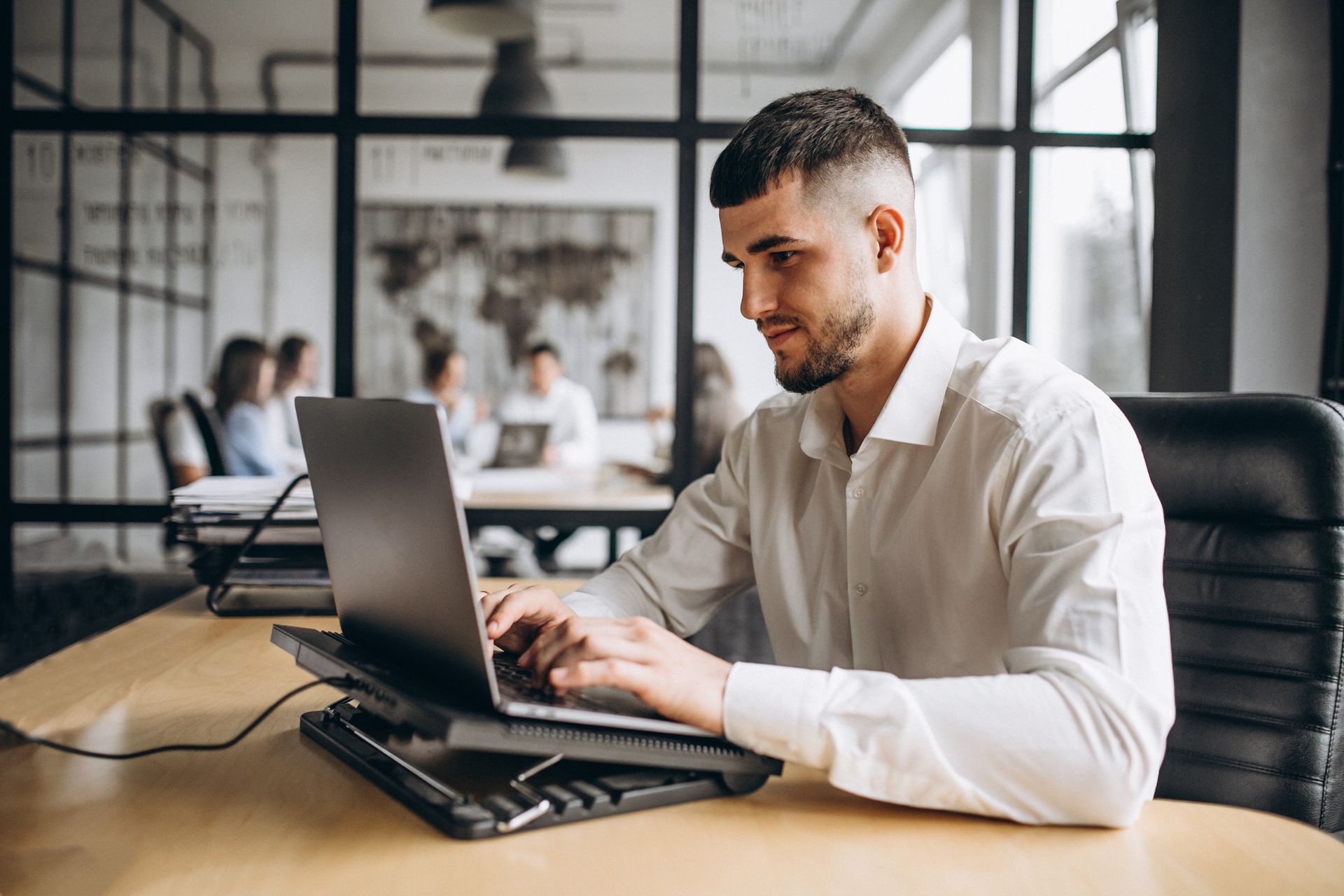 Man typing on a laptop at a conference table in a modern office, with a notebook beside him.