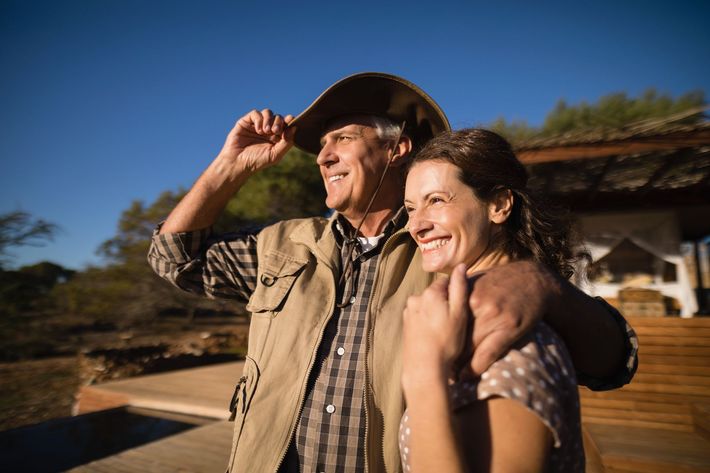 Smiling couple outdoors, man in cowboy hat with arm around woman, standing by a rustic building at sunset