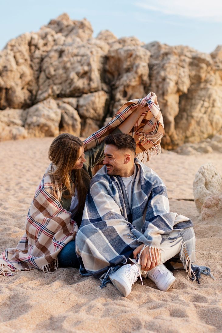 Two people sitting on sand by rocky cliffs, wrapped in patterned blankets in a breezy outdoor setting