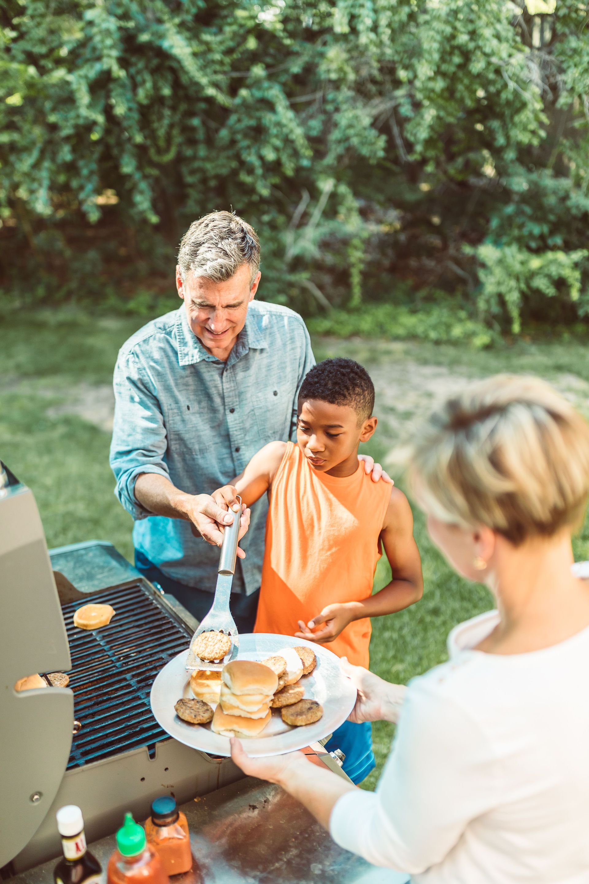 People passing a plate of burgers and buns at an outdoor picnic table.