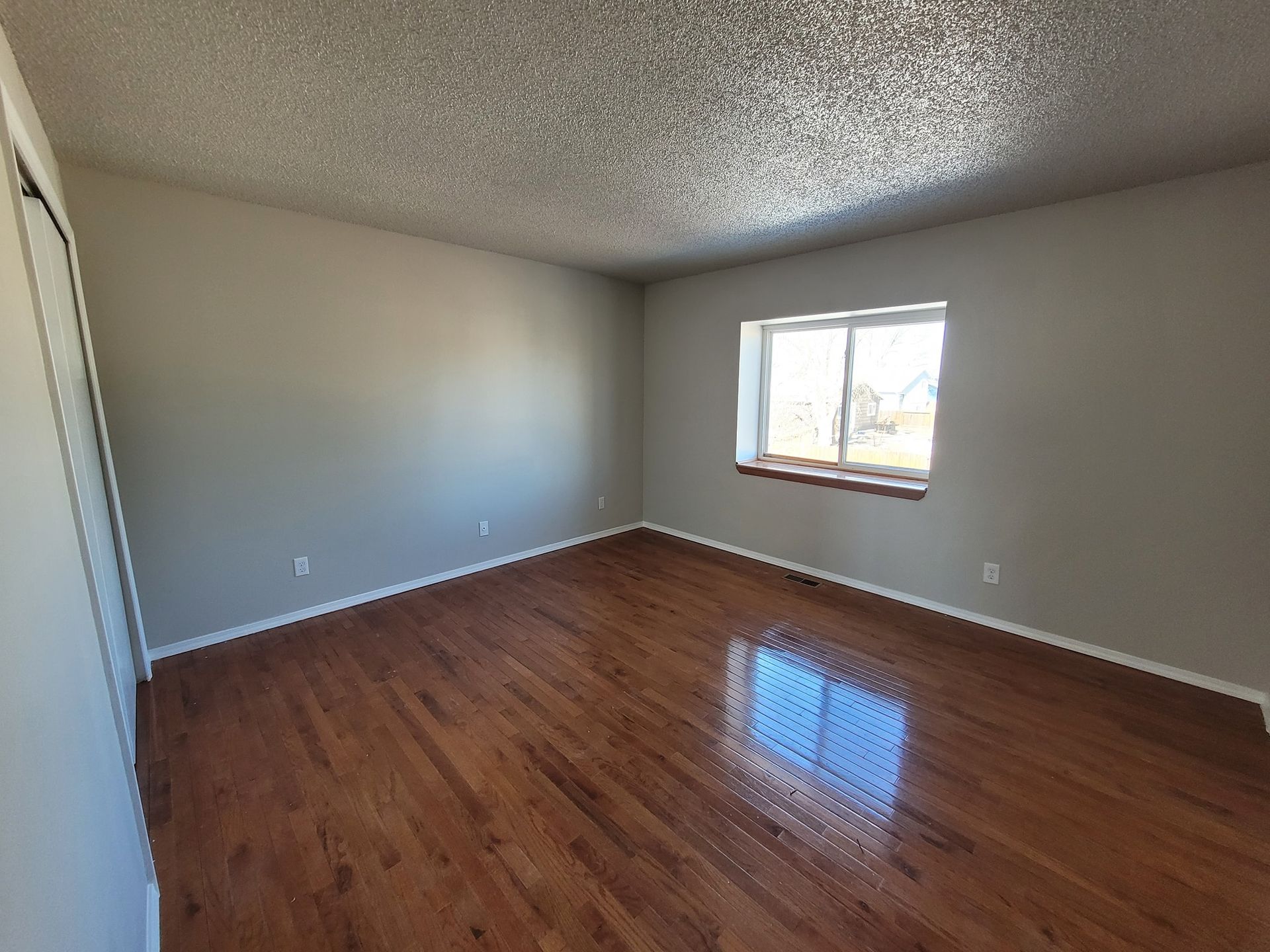 A wide shot of an empty, neutral-toned room with shiny wood floors and a single window on the far wall.