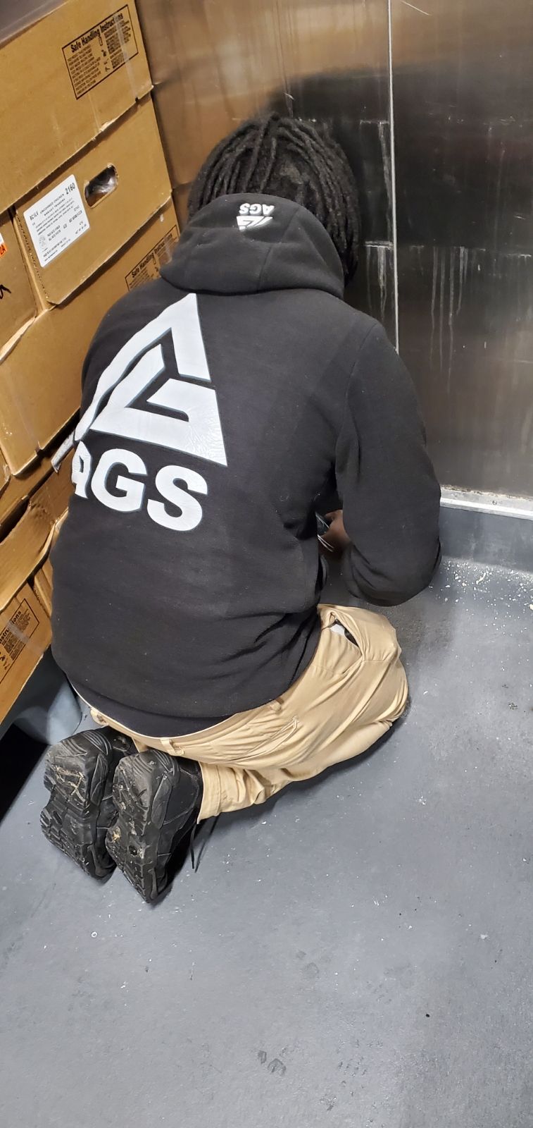 A person wearing a black hoodie and tan pants kneeling on a grey floor next to stacks of cardboard boxes.