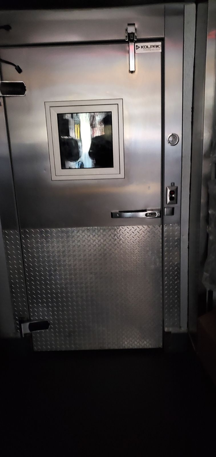 A stainless-steel walk-in cooler door with a square viewing window and diamond plate metal covering the lower portion.