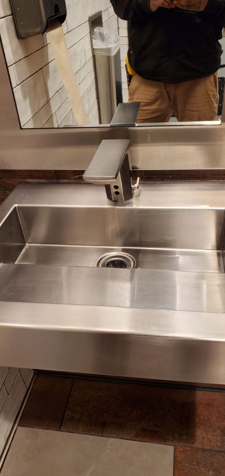 A stainless steel sink and faucet in a restroom with white subway tiles and a paper towel dispenser.