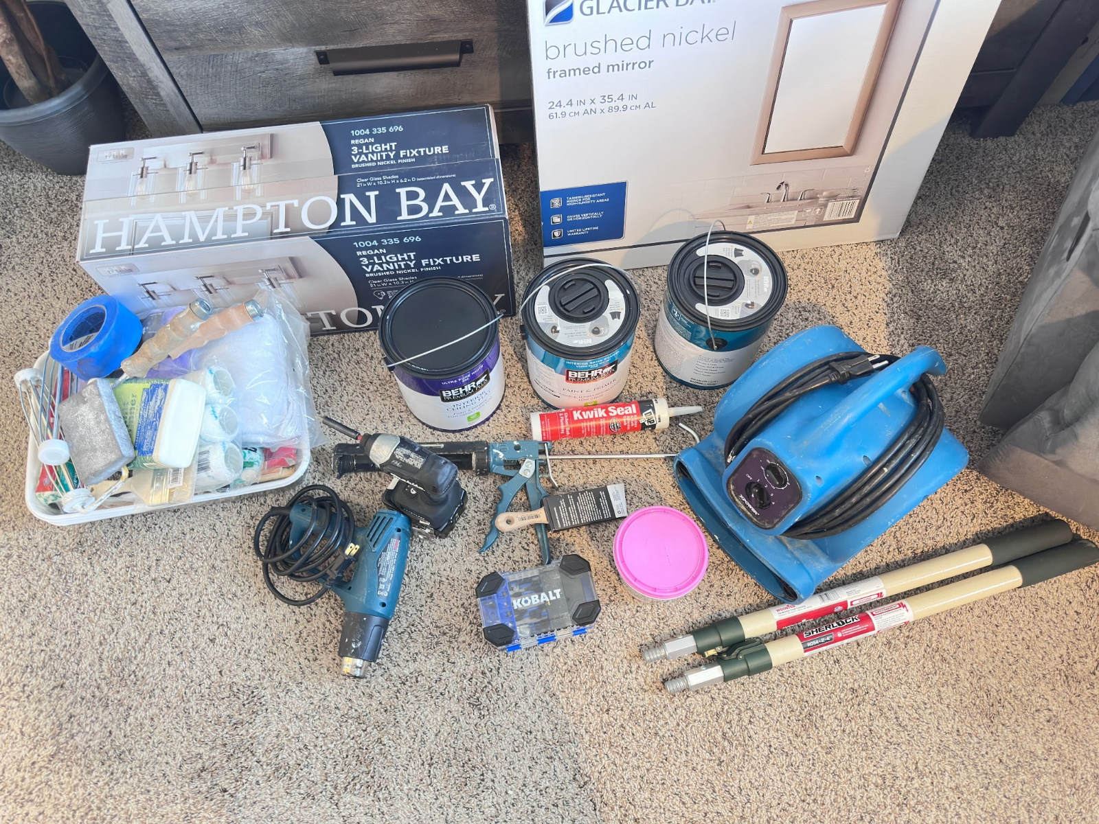 Assorted painting supplies on carpet, including paint cans, tools, rollers, and tape, with a Hampton Bay paint box.