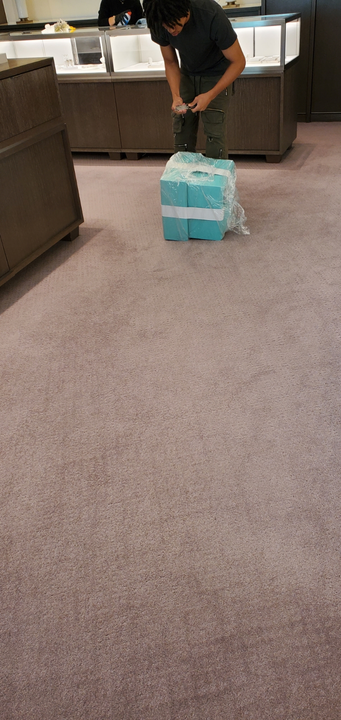 Person unwraps a large box on a carpeted floor in a store. The box is wrapped in blue and clear plastic.