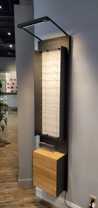 A wall-mounted display unit for eyeglasses featuring a lighted vertical rack above a wooden storage cabinet.