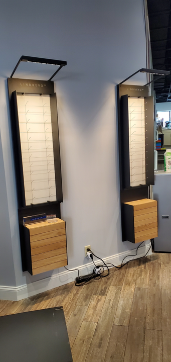 Two wall-mounted display units with illuminated shelves for eyewear and light wood storage cabinets below on a light wall.