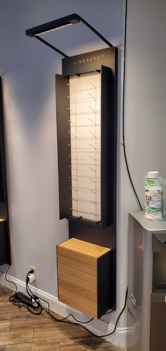 A vertical, wall-mounted display unit for eyeglasses with a lighted, rack-style backboard and a wooden base cabinet.