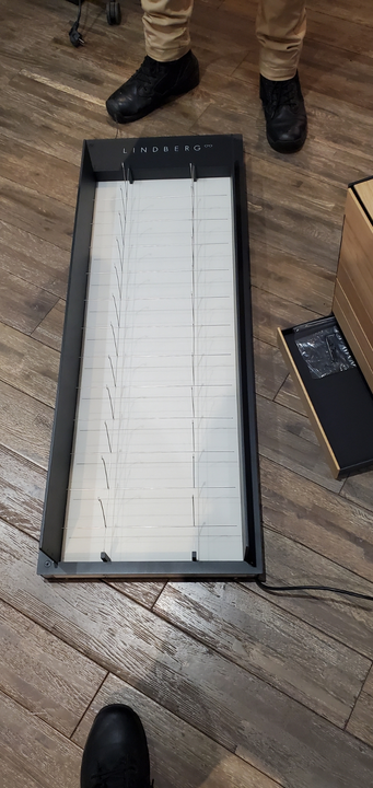 A long, black rectangular display tray with white adjustable dividers sits on a wooden floor near a person’s feet.