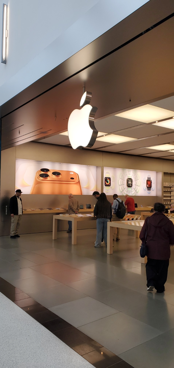 An Apple Store interior with a glowing logo, display tables featuring devices, and people browsing the merchandise.