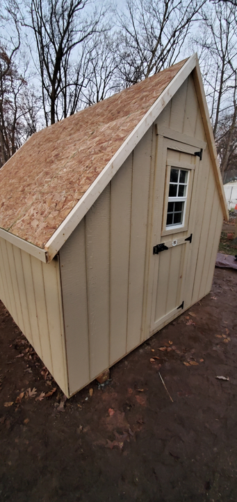 A tan, wooden storage shed with a steep roof and a small window on its front door, set in a wooded, outdoor area.
