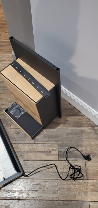 A black and light wood heater stands on a wooden floor next to a wall, with an electrical power cord extending to the side.