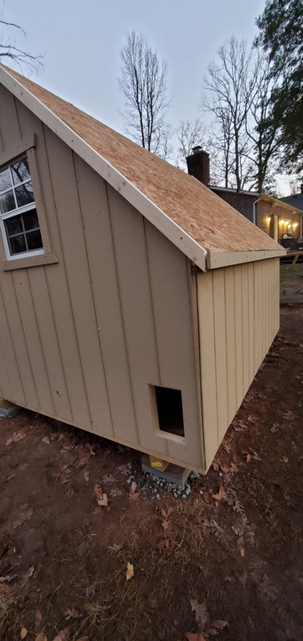 A tan, wood-paneled shed with an unfinished roof and a small square access door at the base, set on a dirt ground.