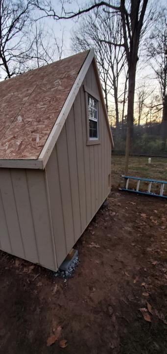 A tan shed with a window under construction, viewed from the side on a dirt lot with trees in the background.