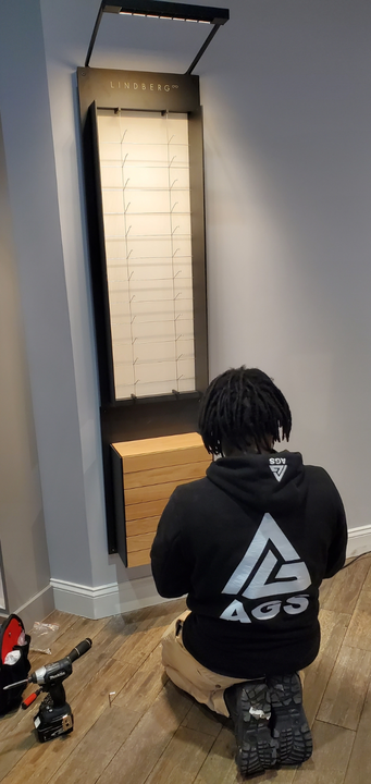 A person kneels while installing a wall-mounted display unit with shelves and lighting in a room with wood-look flooring.