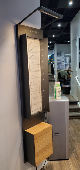 A tall, black wall-mounted display rack for eyeglasses with an integrated light, sitting above a wooden storage cabinet.