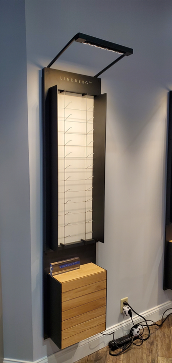 A wall-mounted eyeglass display with a white vertical panel, metal shelving, an overhead light, and a wooden base unit.