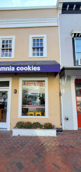 A storefront for Insomnia Cookies on a brick sidewalk, featuring a purple awning, large windows, and small shrubs.