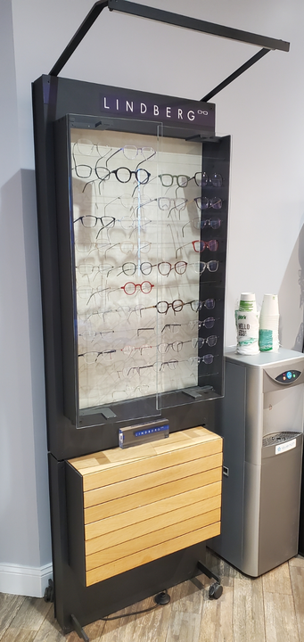 A black, floor-standing display rack for Lindberg eyewear featuring rows of glasses, located next to a water cooler.