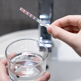 Person testing water with a strip, holding a glass and in front of a faucet.