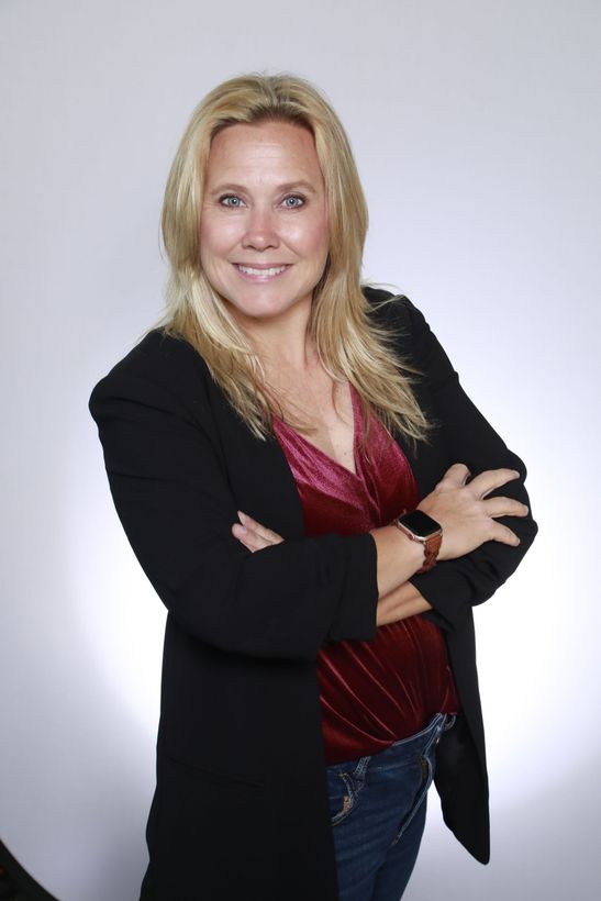 A woman wearing a black jacket and a red top is standing with her arms crossed.