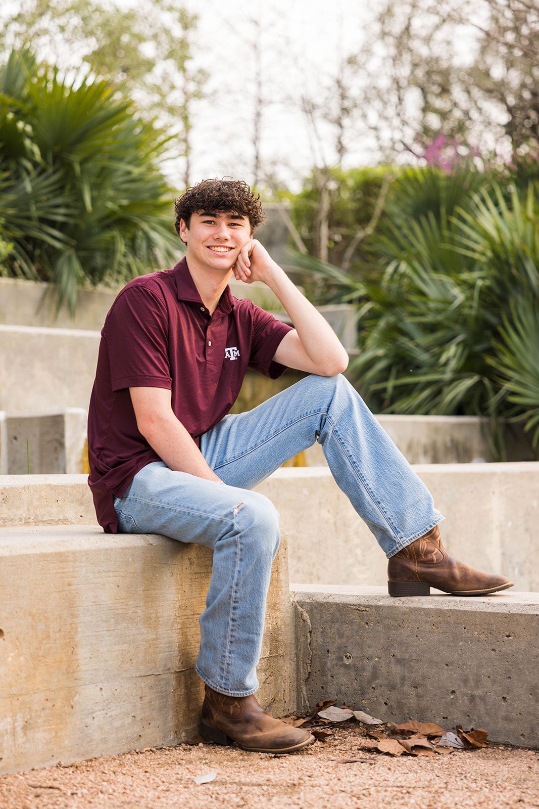 A person in a maroon polo and jeans sits on concrete steps in a park, smiling and resting their chin on their hand.