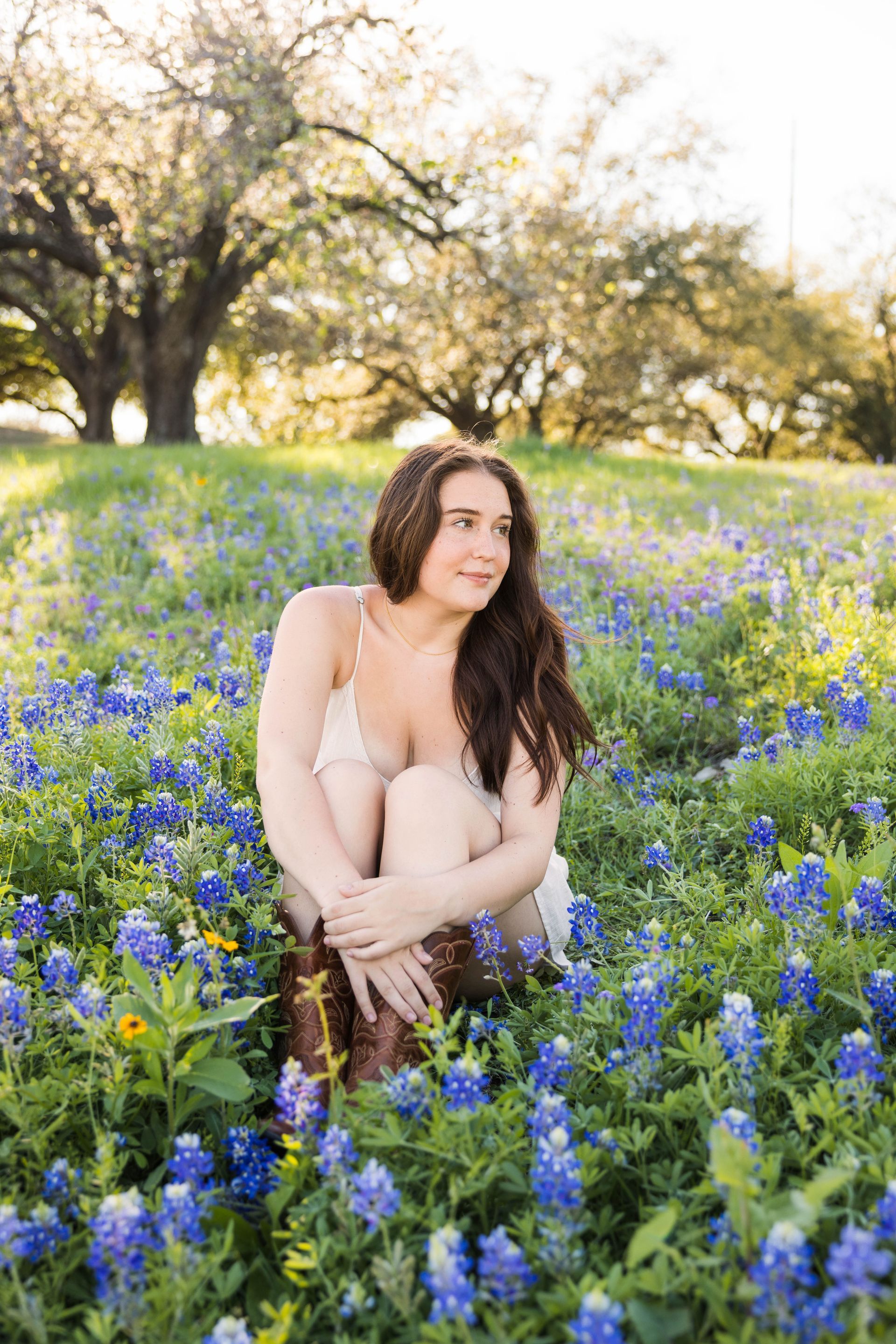 A person sitting in a field of bluebonnets under trees, looking off to the side with a calm expression.