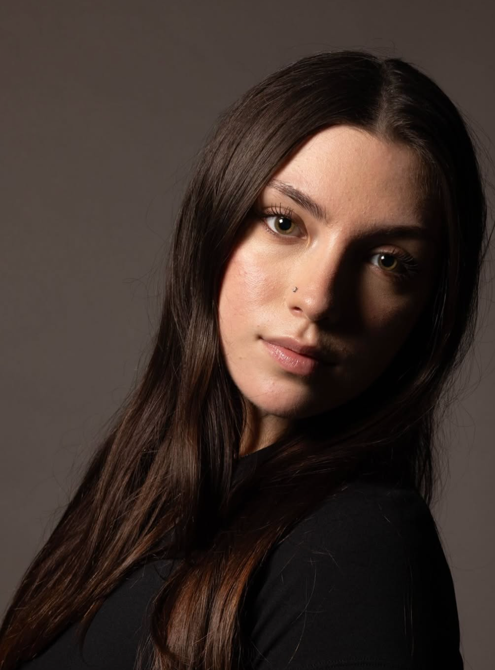 A person with long brown hair posing against a neutral gray background with dramatic side lighting.