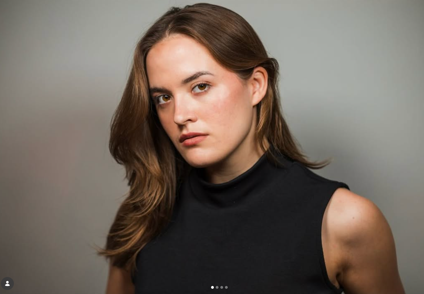 A person with long brown hair, wearing a black sleeveless turtleneck, looks directly at the camera against a gray background.