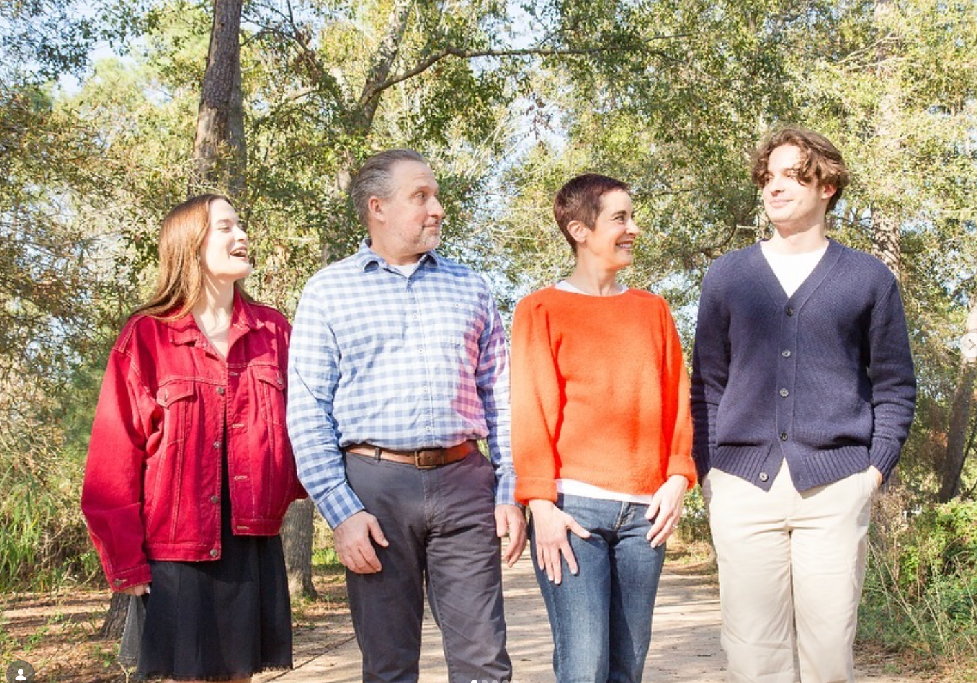 Four people walk together on a sunny, tree-lined path, smiling and looking toward each other in casual outdoor attire.