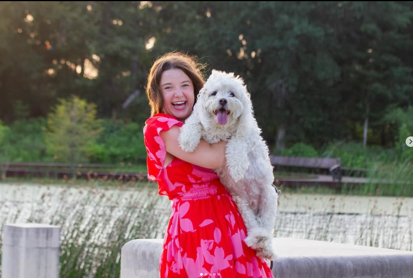 A person in a bright pink and red dress smiling while holding a fluffy white dog outdoors by a lake.