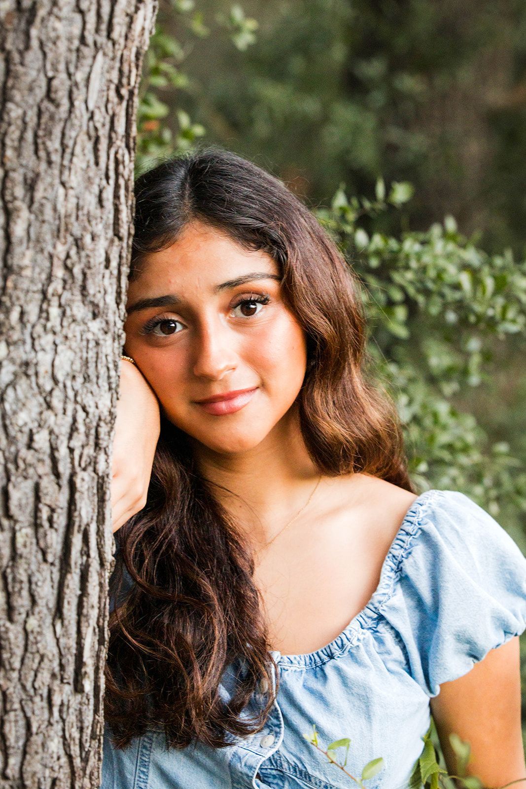 A person with long brown hair, wearing a blue shirt, leans their head against a tree in a nature setting.