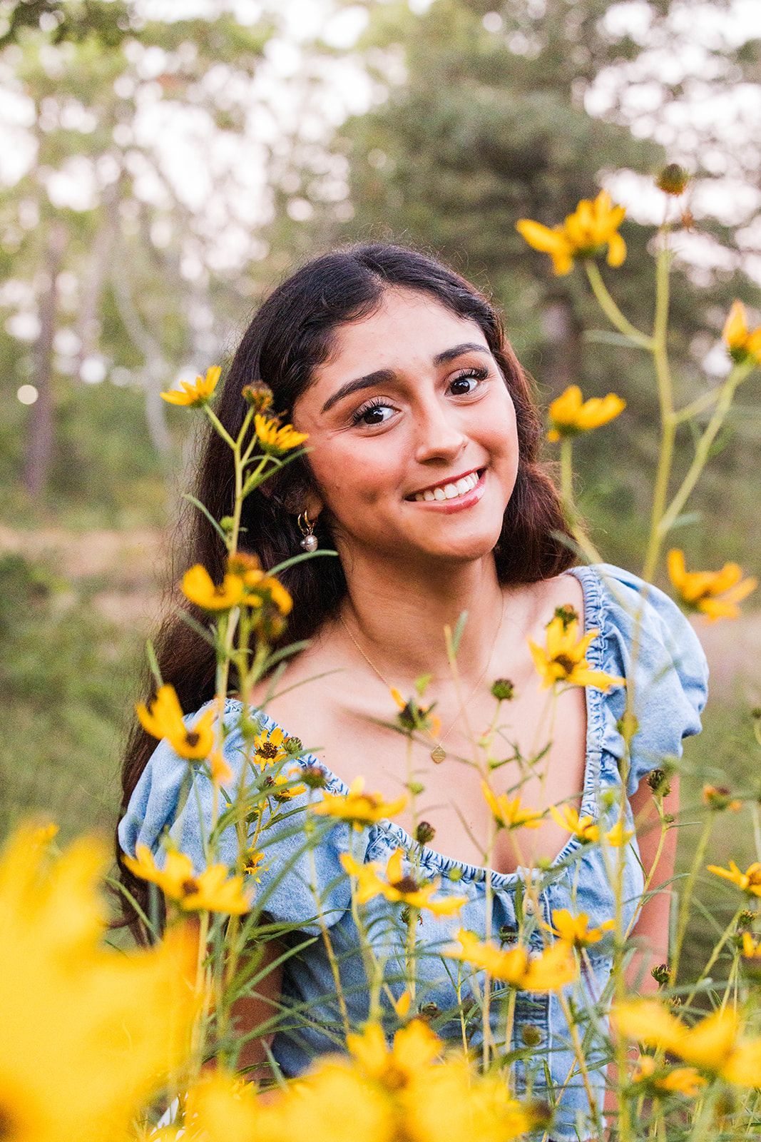 A smiling person in a blue top standing in a field of yellow wildflowers during a sunny day.