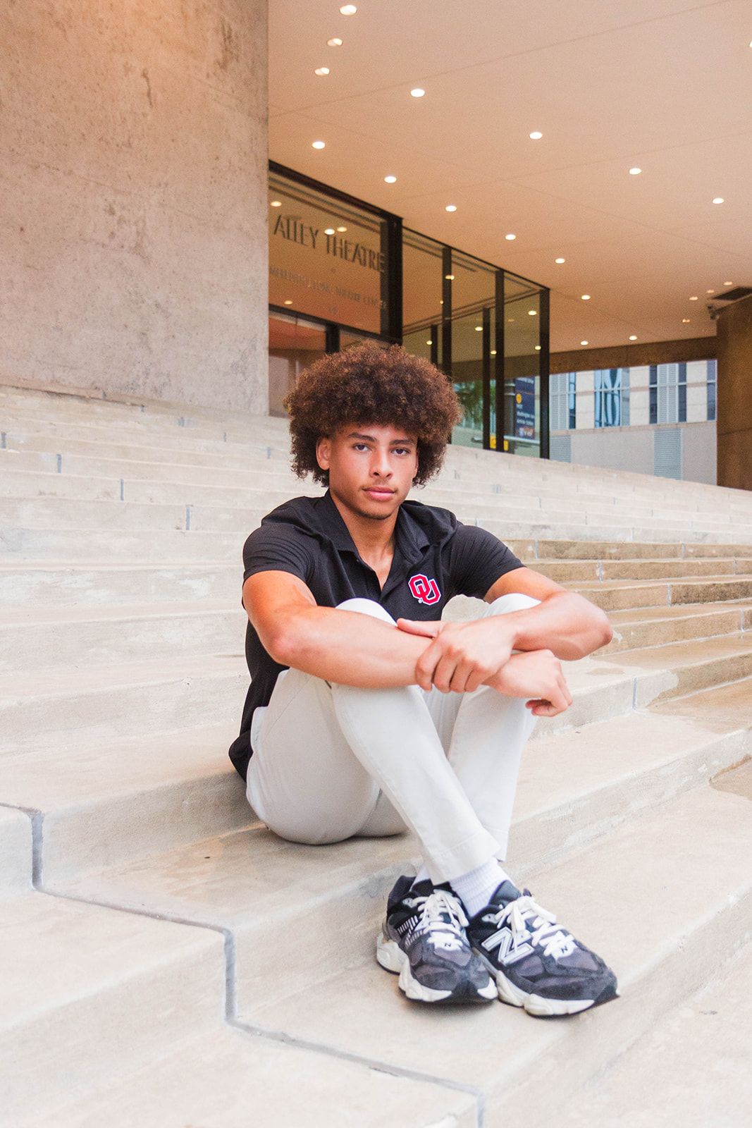 A person with curly hair sits on concrete steps, wearing a black polo shirt, white pants, and athletic sneakers.
