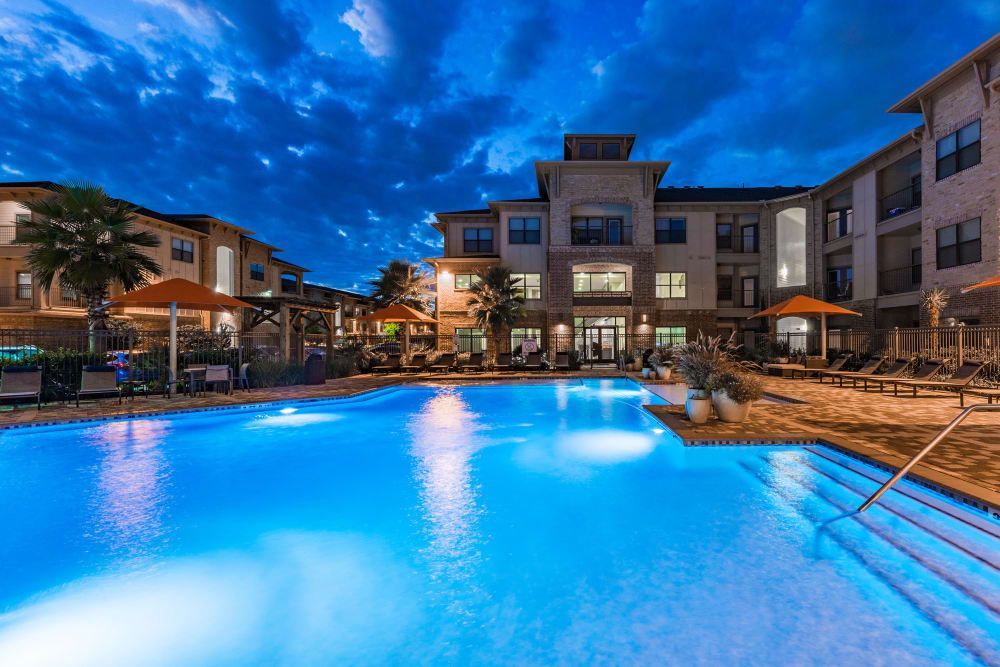 Nighttime view of a large swimming pool in front of a multi-story apartment building with lights reflecting on the water at Marquis Seven Meadows, offering apartments for rent in Katy ISD.