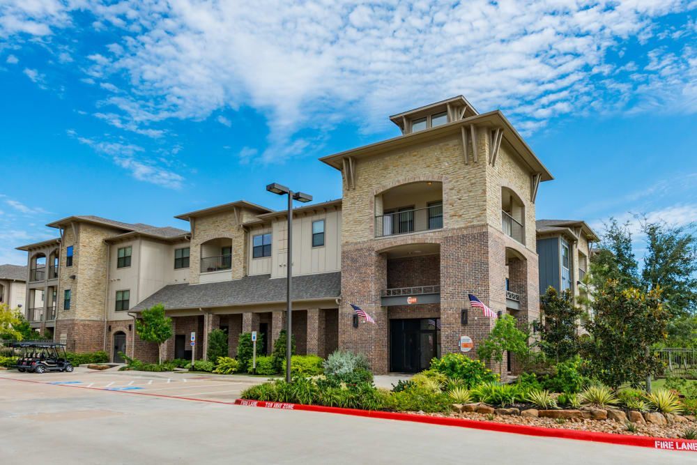 Apartment building with beige and brick facade, blue sky with clouds, landscaping in front at Marquis Seven Meadows, offering apartments for rent in Katy ISD.