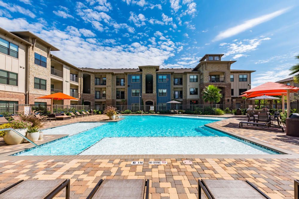 Pool and apartment building exterior at Marquis Seven Meadows, offering apartments for rent in Katy ISD. Blue water, sunny sky. Lounge chairs, orange umbrellas.