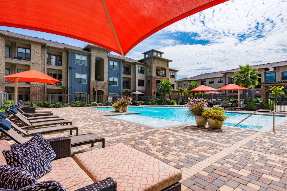 Pool area with lounge chairs, orange umbrellas, and apartment buildings on a sunny day at Marquis Seven Meadows, offering apartments for rent in Katy ISD.