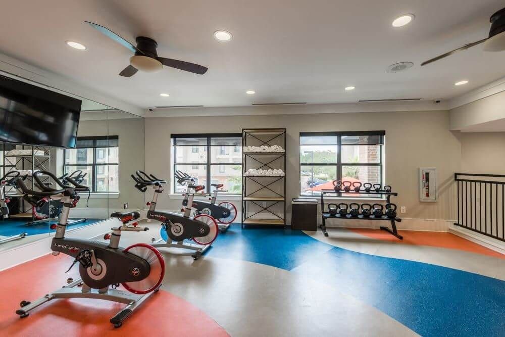 Gym with spin bikes, weights, a large mirror, and ceiling fans at Marquis Seven Meadows, offering apartments in Katy ISD. Blue, orange, and gray floor.
