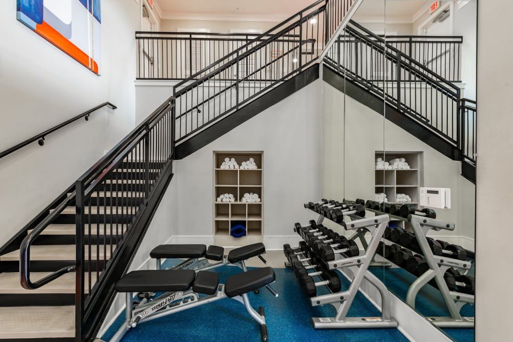Gym with dumbbells, benches, and stairs with metal railings; blue floor, white walls at Marquis Seven Meadows, offering apartments for rent in Katy ISD.