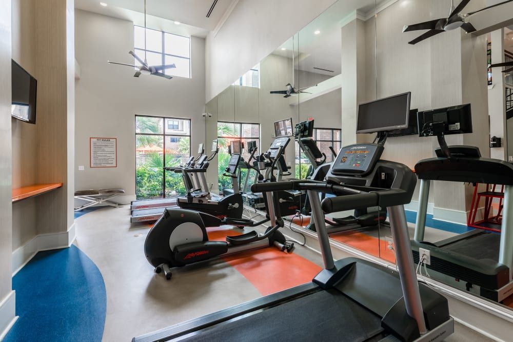 Gym with treadmills, elliptical, weights, and mirrors at Marquis Seven Meadows, offering apartments for rent in Katy ISD. Blue, orange, and gray floors. High ceilings.