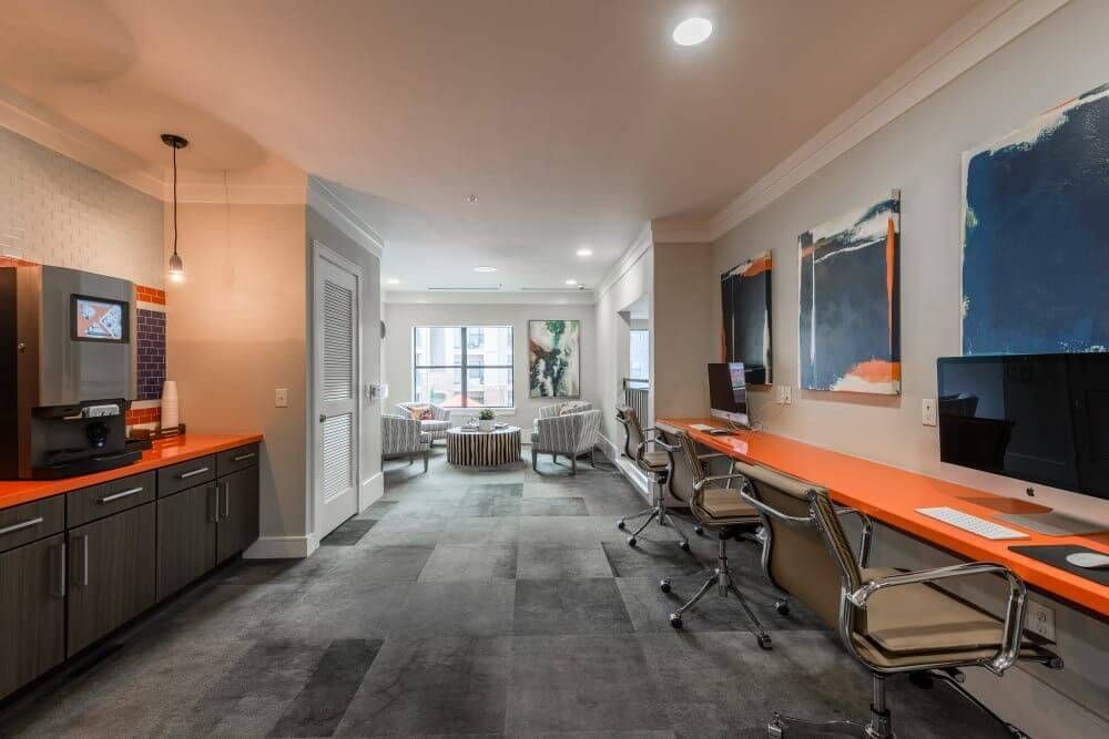 Interior of a modern office space with computer desks, coffee bar, and lounge seating at Marquis Seven Meadows, offering apartments in Katy ISD. Orange and gray tones.