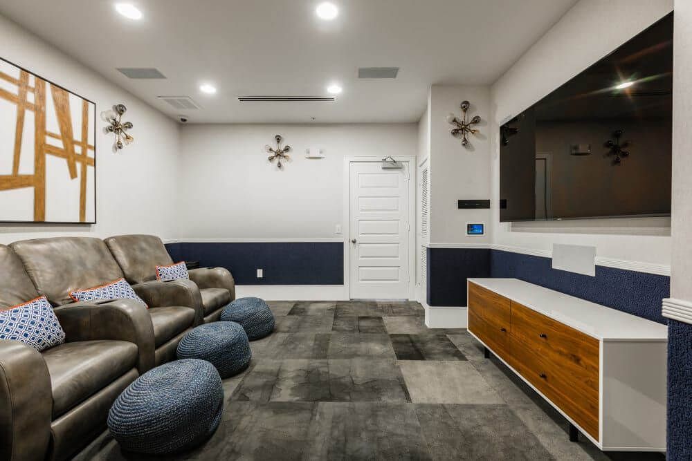 A community theater with leather recliners, a large TV, blue and gold accents, and patterned carpet at Marquis Seven Meadows, offering apartments in Katy ISD.
