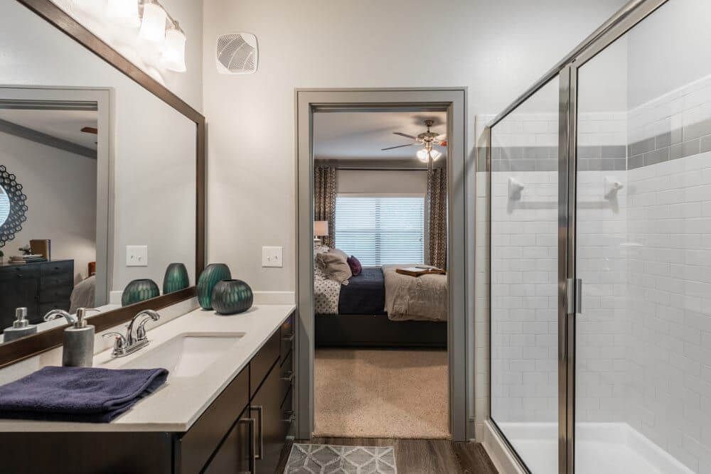 Bathroom with a vanity, shower, and doorway to a bedroom. Dark wood cabinets, white countertop, and gray walls at Marquis Seven Meadows, offering apartments in Katy, TX.