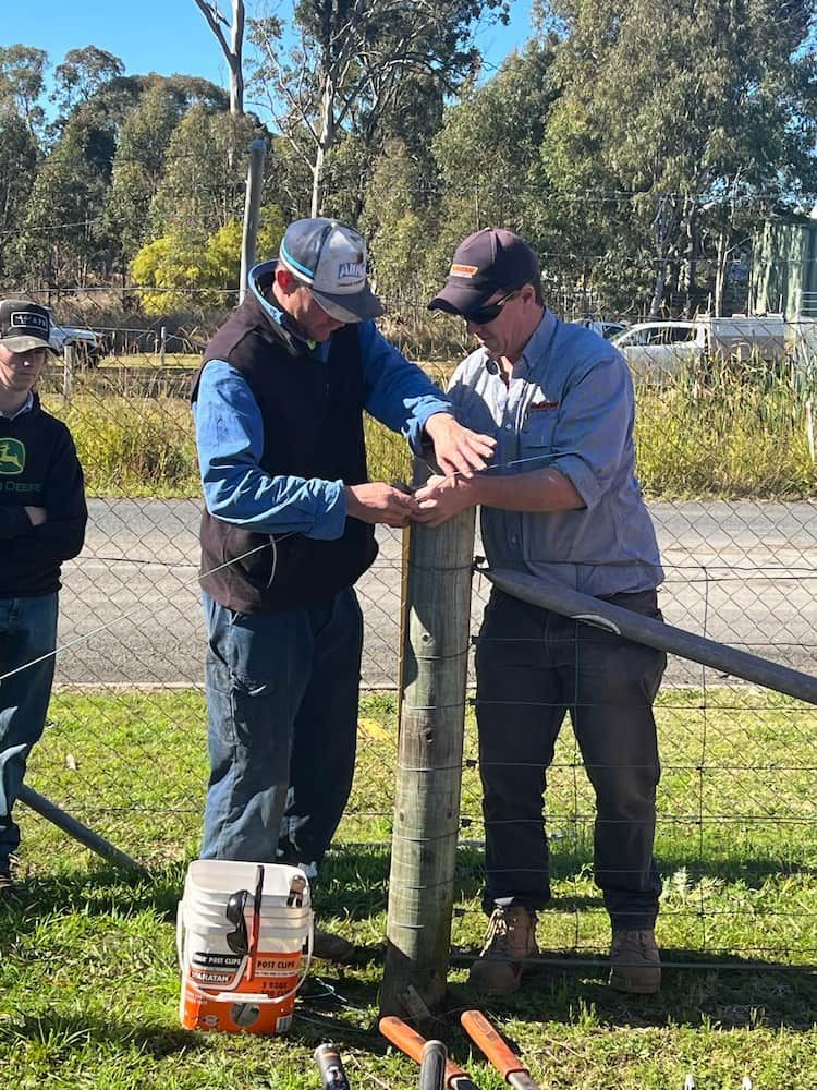 Two Men Work on Fence Post in Grassy Outdoor Area — Gloucester Rural Supplies in Bulahdelah, NSW