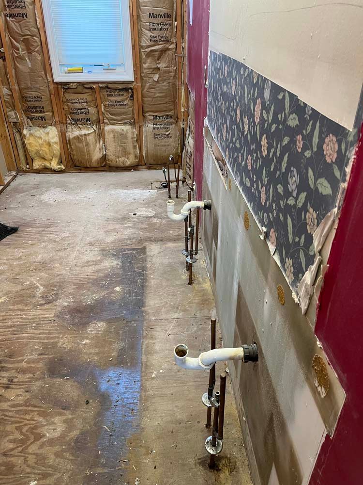 Bathroom under renovation with exposed wall studs, plumbing pipes, and torn flooring.