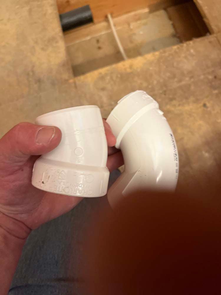 Hand holding two white PVC pipe elbow fittings over a tiled floor and sink area
