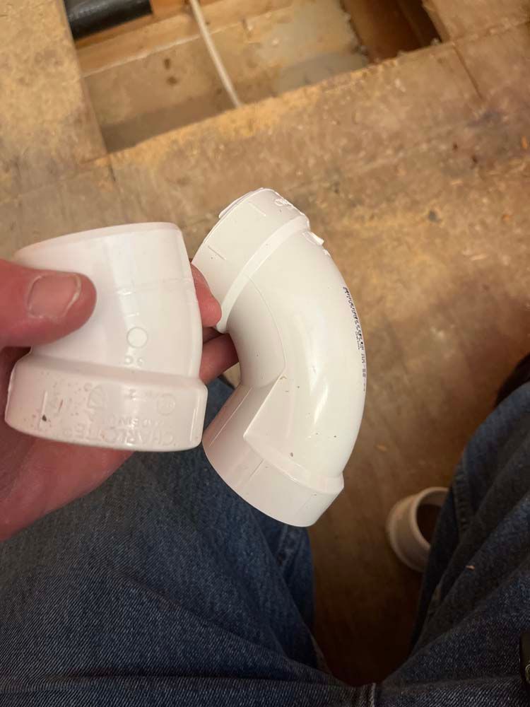 Hand holding a broken white PVC pipe fitting over jeans on a floor.