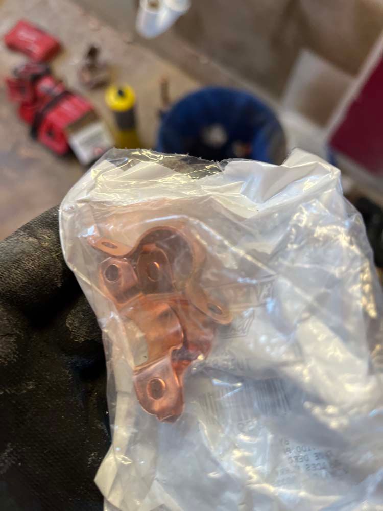 Hand in black glove holding a small copper automotive part in a clear plastic bag, with tools blurred in background